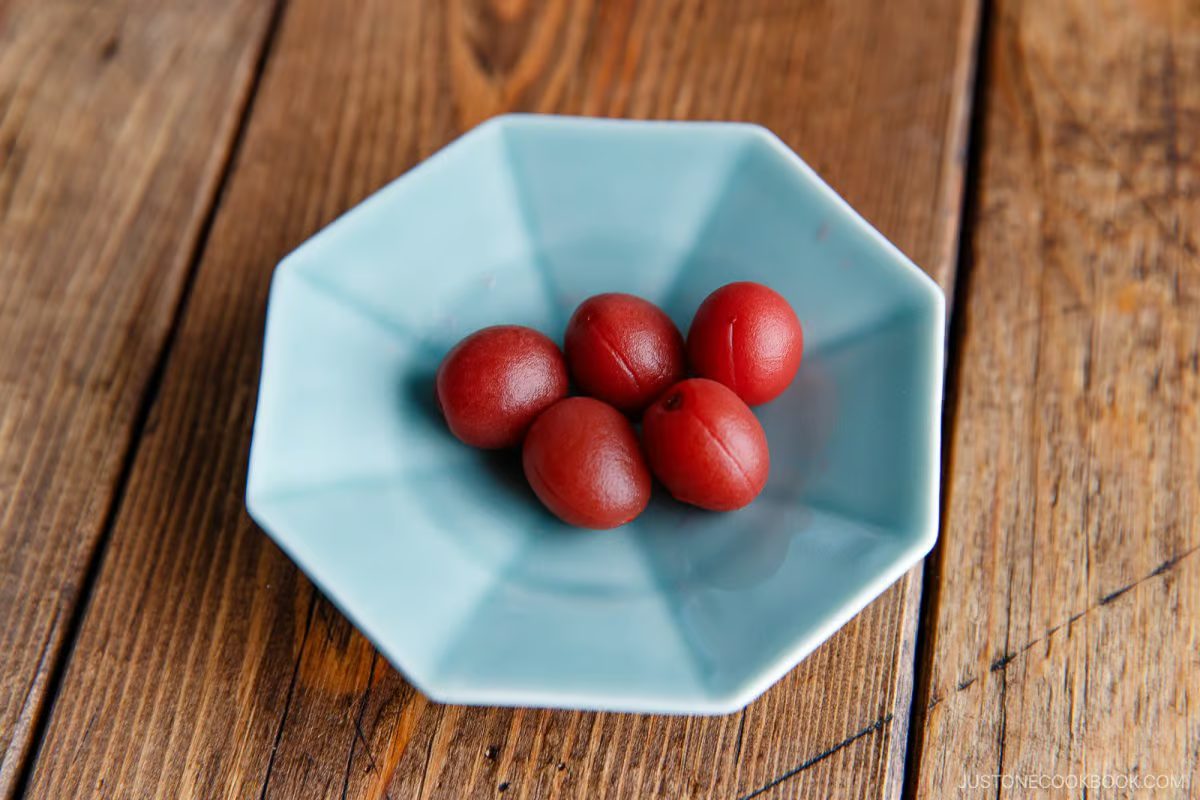 Five pickled plums (umeboshi), reminiscent of those featured in the Umeboshi Onigiri (Plum Rice Ball) from Midnight Diner, rest on a light blue octagonal plate, placed on a wooden table.