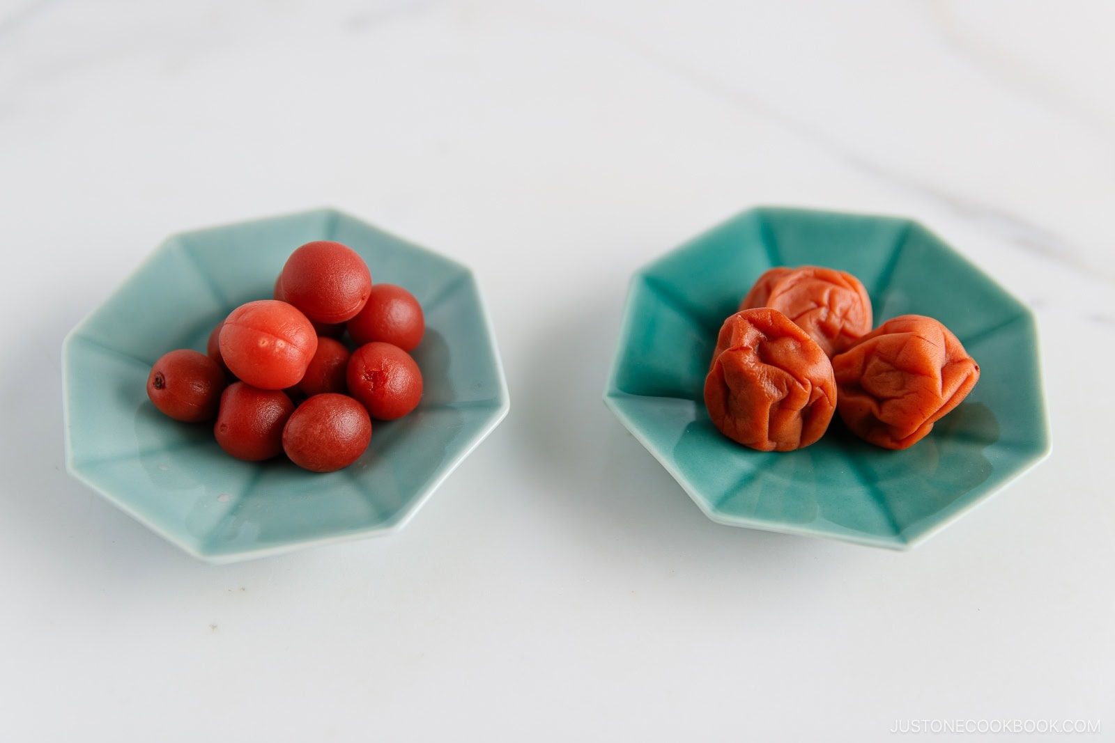Two teal dishes on a white surface; the left dish holds small red pickled plums, while the right dish contains larger, wrinkled, reddish-brown pickled plums—ingredients often featured in dishes like Umeboshi Onigiri (Plum Rice Ball) from Midnight Diner.