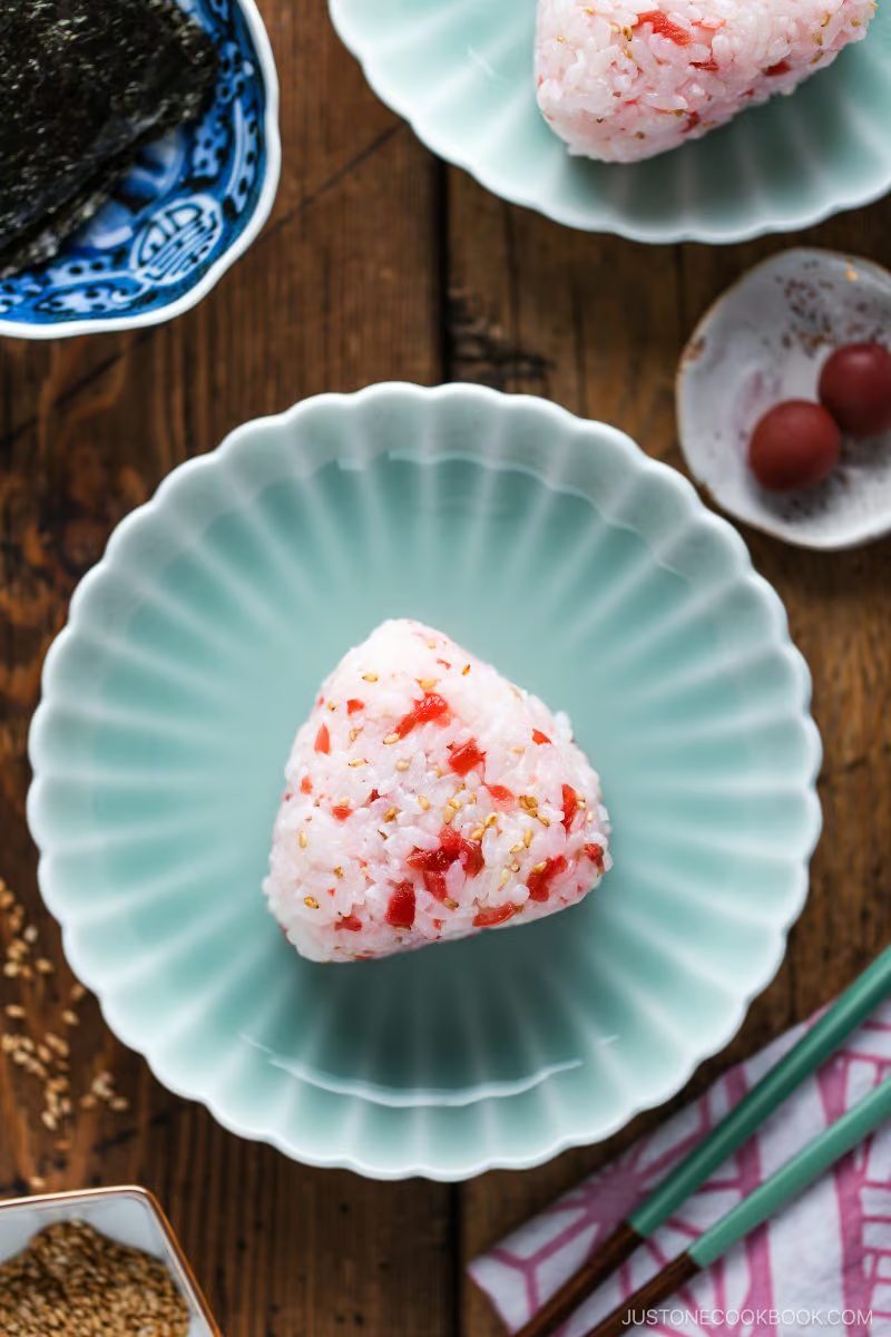 A triangular rice ball with red bits, possibly pickled plum, sits on a pale green scalloped plate. This Umeboshi Onigiri (Plum Rice Ball), reminiscent of those from Midnight Diner, is accompanied by chopsticks, sesame seeds, seaweed, and extra pickled plums on a wooden table.