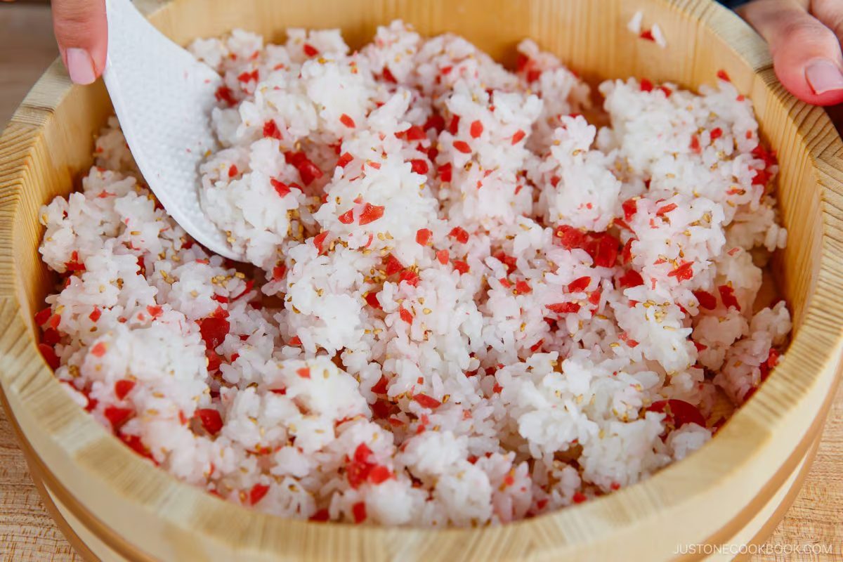 A wooden bowl filled with white rice mixed with small red bits—reminiscent of the Umeboshi Onigiri (Plum Rice Ball) from Midnight Diner—is being stirred with a white rice paddle by a hand.