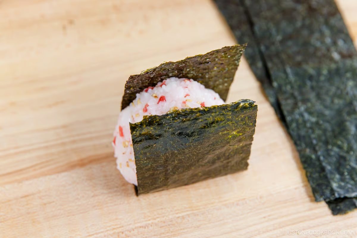 A classic Umeboshi Onigiri (Plum Rice Ball), inspired by Midnight Diner, sits on a wooden surface wrapped with two rectangular sheets of nori seaweed, with extra nori strips nearby.