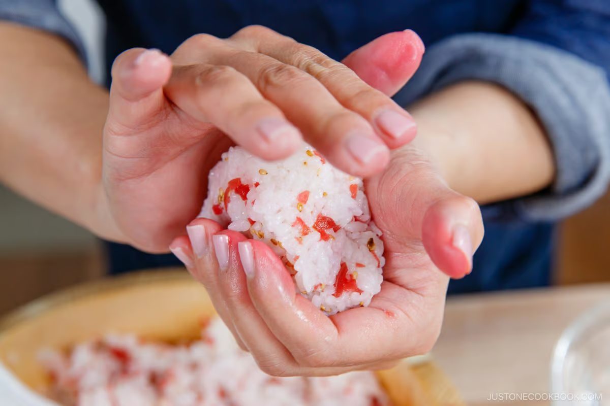 A person shapes a Umeboshi Onigiri (Plum Rice Ball) inspired by Midnight Diner, mixing red ingredients and sesame seeds into the rice with their hands, while a tray of similar rice balls sits in the background.