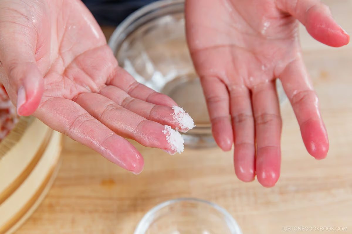 Two hands with slightly wet fingers hold a small amount of salt between the thumb and fingers over a wooden surface, with a glass bowl in the background—a simple yet essential step in preparing Umeboshi Onigiri (Plum Rice Ball) as featured in Midnight Diner.