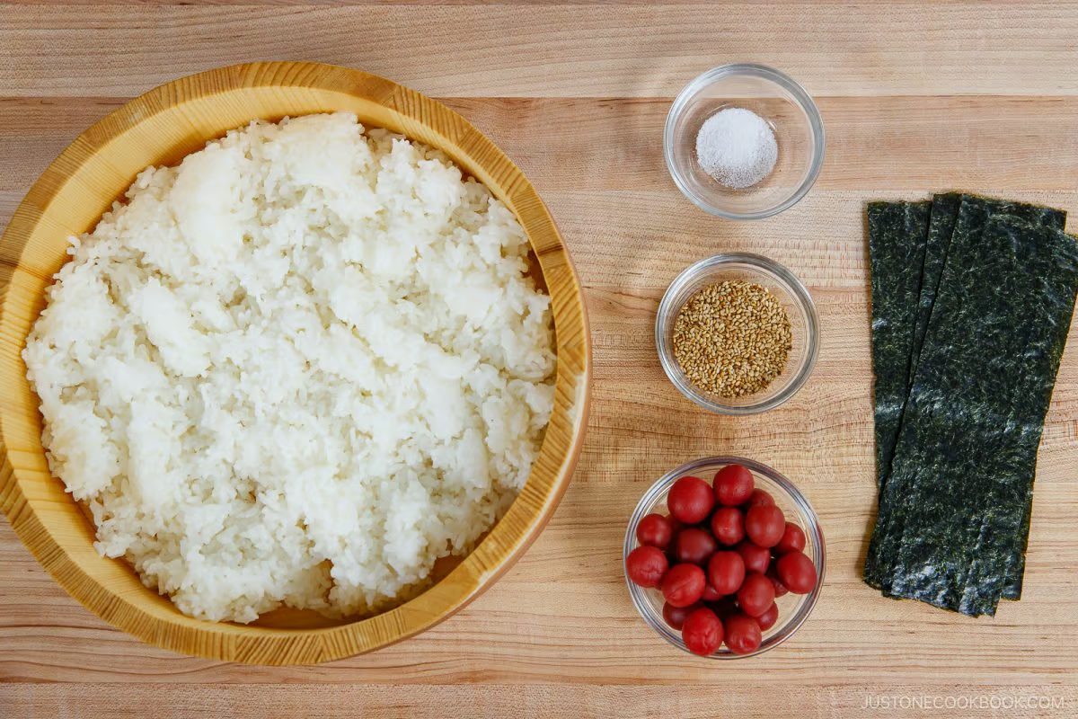 A wooden bowl of white rice, a small bowl of salt, a bowl of sesame seeds, a bowl of red pickled plums, and seaweed sheets are arranged on a wooden surface—perfect ingredients for crafting Umeboshi Onigiri (Plum Rice Ball) inspired by Midnight Diner.