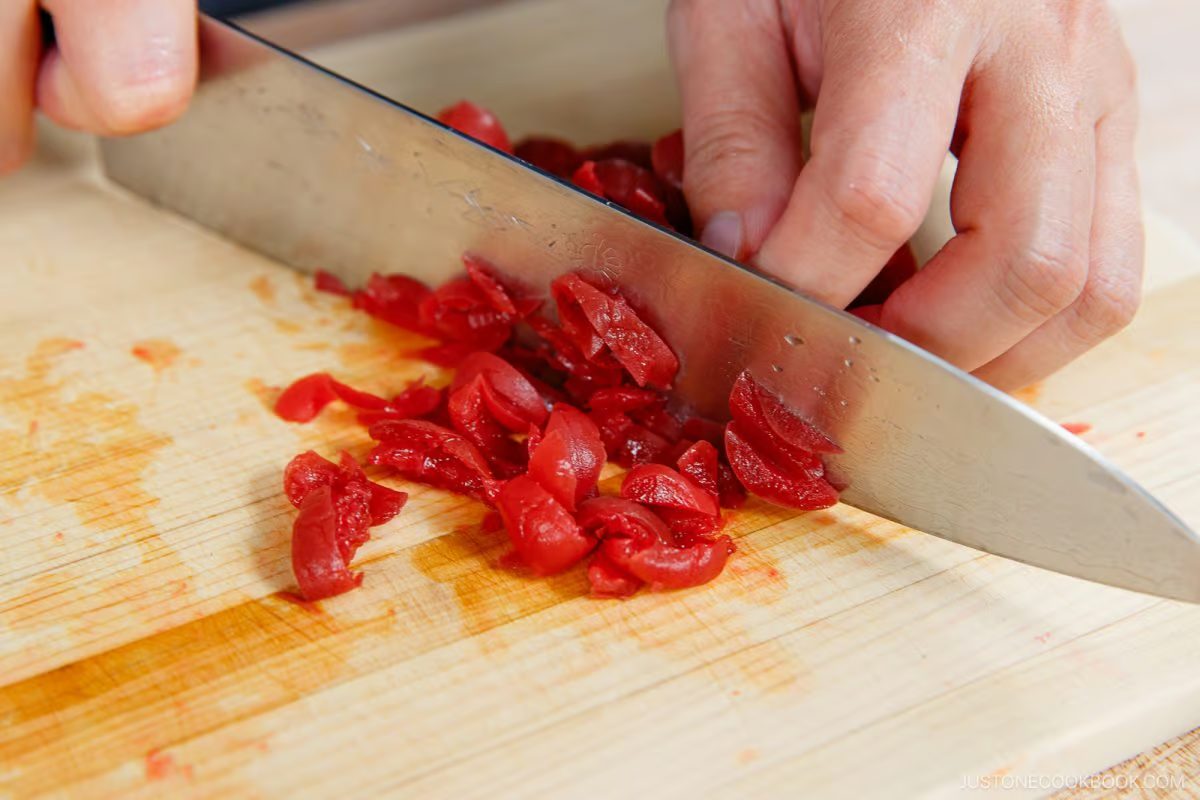 A person finely chops red pickled vegetables, perhaps preparing ingredients for Umeboshi Onigiri (Plum Rice Ball) as seen in Midnight Diner, on a wooden cutting board with a large kitchen knife.