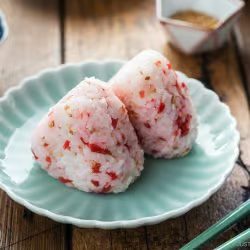 Two Umeboshi Onigiri (Plum Rice Balls), inspired by Midnight Diner, with red seasoning sit on a pale-green plate, accompanied by green chopsticks. A wooden table, pickled garnish, water glass, and small dishes are visible in the background.