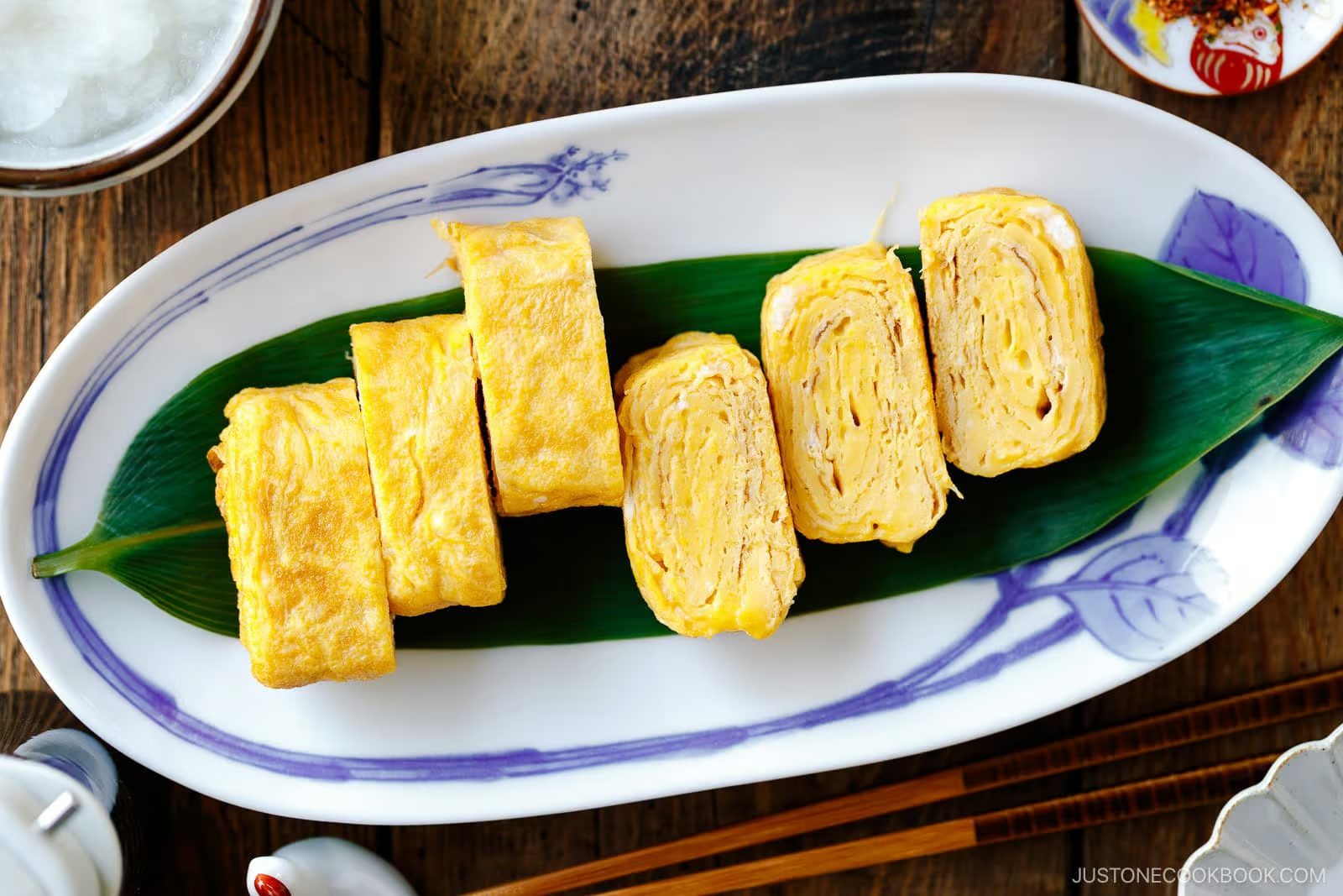 Five pieces of Dashimaki Tamago (Japanese Dashi Rolled Omelette) are neatly arranged on a green leaf atop a rectangular white plate with blue accents, set on a wooden table with various small dishes nearby.
