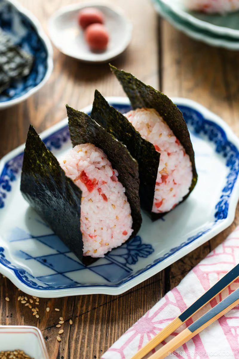Two Umeboshi Onigiri (Plum Rice Ball) as featured in Midnight Diner, wrapped in seaweed and filled with tangy pink plum, sit on a blue and white plate surrounded by Japanese tableware and garnishes on a wooden surface.
